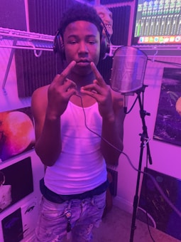 A person wearing headphones and a white tank top stands in a music studio. They are gesturing with their hands, positioned in front of a microphone with a pop filter. The room has purple lighting and is surrounded by soundproof panels and various artworks. A computer screen with audio mixing software is visible in the background.