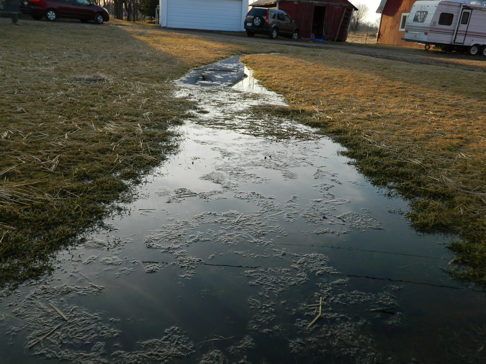 A long strip of standing water lying across flat grass in front of a parked travel trailer and two cars