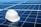 A white hard hat rests on a large array of blue solar panels under a clear sky. The panels are aligned in an orderly fashion, reflecting sunlight and emphasizing a clean energy theme.