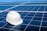 A white hard hat rests on a large array of blue solar panels under a clear sky. The panels are aligned in an orderly fashion, reflecting sunlight and emphasizing a clean energy theme.