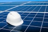 A white hard hat rests on a large array of blue solar panels under a clear sky. The panels are aligned in an orderly fashion, reflecting sunlight and emphasizing a clean energy theme.