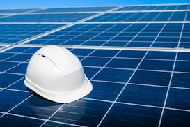 A white hard hat rests on a large array of blue solar panels under a clear sky. The panels are aligned in an orderly fashion, reflecting sunlight and emphasizing a clean energy theme.