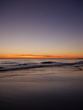 The image captures a serene sunset over a calm ocean. Gentle waves roll towards a sandy shore, with the sky transitioning from deep blue at the top to a warm orange near the horizon. A small airplane can be seen silhouetted against the colorful sky, adding a sense of scale and depth.
