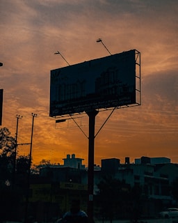 A large billboard is silhouetted against a vibrant orange and yellow sunset sky, featuring a building advertisement. Power lines and the outlines of buildings are visible in the foreground. The overall scene is urban with a tranquil atmosphere.