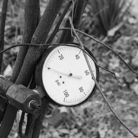 A black and white close-up of a pressure gauge attached to metal pipes. The gauge displays measurements in psi, ranging from 0 to 100. Several cables or strings are wrapped around the pipes, and the background appears to be an outdoor setting with blurred foliage.