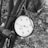 A black and white close-up of a pressure gauge attached to metal pipes. The gauge displays measurements in psi, ranging from 0 to 100. Several cables or strings are wrapped around the pipes, and the background appears to be an outdoor setting with blurred foliage.