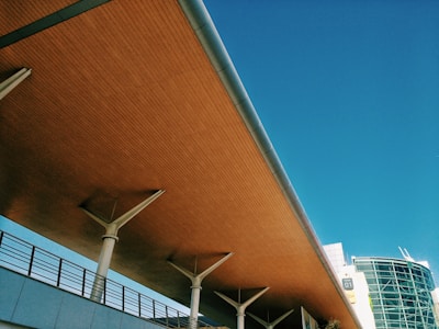 A modern architectural structure with a wooden ceiling and metal supports stretches at an angle against a clear blue sky. A section of a glass-fronted building is visible in the background, along with a street light.