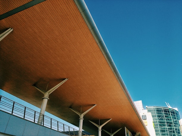 A modern architectural structure with a wooden ceiling and metal supports stretches at an angle against a clear blue sky. A section of a glass-fronted building is visible in the background, along with a street light.