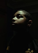 A dramatic profile portrait of a person with shadows cast across their face. The lighting highlights the contours of the face, creating an intense and introspective mood. The background is dark, enhancing the focus on the person's facial features.