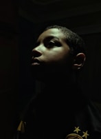 A dramatic profile portrait of a person with shadows cast across their face. The lighting highlights the contours of the face, creating an intense and introspective mood. The background is dark, enhancing the focus on the person's facial features.