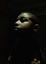 A dramatic profile portrait of a person with shadows cast across their face. The lighting highlights the contours of the face, creating an intense and introspective mood. The background is dark, enhancing the focus on the person's facial features.