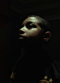 A dramatic profile portrait of a person with shadows cast across their face. The lighting highlights the contours of the face, creating an intense and introspective mood. The background is dark, enhancing the focus on the person's facial features.
