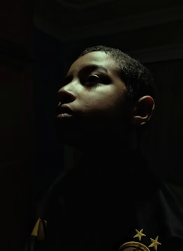 A dramatic profile portrait of a person with shadows cast across their face. The lighting highlights the contours of the face, creating an intense and introspective mood. The background is dark, enhancing the focus on the person's facial features.