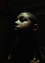 A dramatic profile portrait of a person with shadows cast across their face. The lighting highlights the contours of the face, creating an intense and introspective mood. The background is dark, enhancing the focus on the person's facial features.