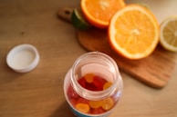 A jar filled with colorful gummy vitamins is placed on a wooden table. The jar is open, with the cap lying beside it. In the background, there is a wooden cutting board with halved oranges, a lemon, and a lime, adding a vibrant touch to the scene.