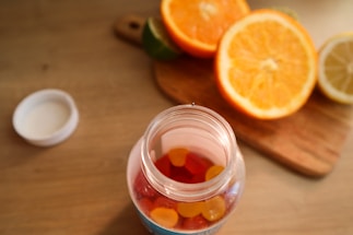 A jar filled with colorful gummy vitamins is placed on a wooden table. The jar is open, with the cap lying beside it. In the background, there is a wooden cutting board with halved oranges, a lemon, and a lime, adding a vibrant touch to the scene.