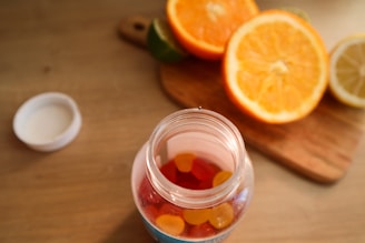 A jar filled with colorful gummy vitamins is placed on a wooden table. The jar is open, with the cap lying beside it. In the background, there is a wooden cutting board with halved oranges, a lemon, and a lime, adding a vibrant touch to the scene.
