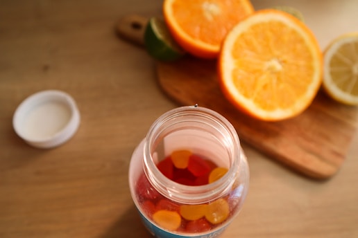 A jar filled with colorful gummy vitamins is placed on a wooden table. The jar is open, with the cap lying beside it. In the background, there is a wooden cutting board with halved oranges, a lemon, and a lime, adding a vibrant touch to the scene.