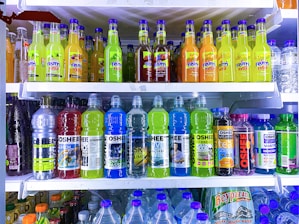 A refrigerator is filled with a variety of colorful bottled drinks. The top shelf displays fruit-flavored beverages with vibrant labels, including green, yellow, and orange colors. The middle shelf features sports and energy drinks in large plastic bottles with labels that use metallic and bright blue colors. The bottom section has a collection of water bottles and other cartons of beverages, showing a mix of branding and flavors.