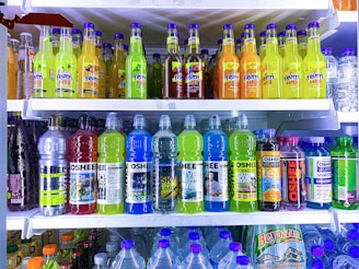 A refrigerator is filled with a variety of colorful bottled drinks. The top shelf displays fruit-flavored beverages with vibrant labels, including green, yellow, and orange colors. The middle shelf features sports and energy drinks in large plastic bottles with labels that use metallic and bright blue colors. The bottom section has a collection of water bottles and other cartons of beverages, showing a mix of branding and flavors.