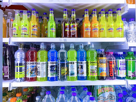A refrigerator is filled with a variety of colorful bottled drinks. The top shelf displays fruit-flavored beverages with vibrant labels, including green, yellow, and orange colors. The middle shelf features sports and energy drinks in large plastic bottles with labels that use metallic and bright blue colors. The bottom section has a collection of water bottles and other cartons of beverages, showing a mix of branding and flavors.
