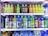 A refrigerator is filled with a variety of colorful bottled drinks. The top shelf displays fruit-flavored beverages with vibrant labels, including green, yellow, and orange colors. The middle shelf features sports and energy drinks in large plastic bottles with labels that use metallic and bright blue colors. The bottom section has a collection of water bottles and other cartons of beverages, showing a mix of branding and flavors.
