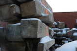 Large wooden beams are stacked in an outdoor industrial area, partially covered with snow. The beams have visible cracks and some red markings on them. A brick building is in the background under a bright sky.