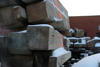 Large wooden beams are stacked in an outdoor industrial area, partially covered with snow. The beams have visible cracks and some red markings on them. A brick building is in the background under a bright sky.
