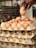 A hand is reaching towards a stack of egg cartons, each filled with brown eggs. The background is slightly blurred, emphasizing the focus on the eggs and hand. The scene appears to be in a market or store setting.