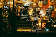 A cluttered shop window filled with a variety of vintage and eclectic items, including lamps, statues, and old mechanical devices. The display is densely packed, creating an antique and nostalgic atmosphere with warm lighting.