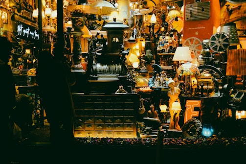A cluttered shop window filled with a variety of vintage and eclectic items, including lamps, statues, and old mechanical devices. The display is densely packed, creating an antique and nostalgic atmosphere with warm lighting.