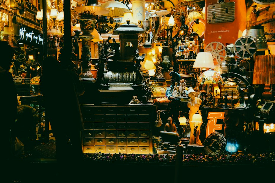 A cluttered shop window filled with a variety of vintage and eclectic items, including lamps, statues, and old mechanical devices. The display is densely packed, creating an antique and nostalgic atmosphere with warm lighting.
