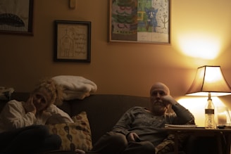 A warmly lit living room with two people sitting on a couch. There are framed artworks on the wall and a table lamp casting a soft glow. One person appears thoughtful or pensive, while the other seems relaxed. A bottle of water and a notebook are on the side table.
