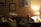 A warmly lit living room with two people sitting on a couch. There are framed artworks on the wall and a table lamp casting a soft glow. One person appears thoughtful or pensive, while the other seems relaxed. A bottle of water and a notebook are on the side table.