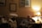 A warmly lit living room with two people sitting on a couch. There are framed artworks on the wall and a table lamp casting a soft glow. One person appears thoughtful or pensive, while the other seems relaxed. A bottle of water and a notebook are on the side table.