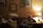 A warmly lit living room with two people sitting on a couch. There are framed artworks on the wall and a table lamp casting a soft glow. One person appears thoughtful or pensive, while the other seems relaxed. A bottle of water and a notebook are on the side table.
