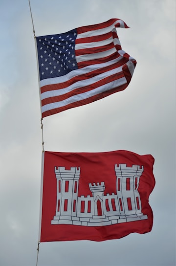 Two flags are flying against a cloudy sky. The upper flag is the United States flag, featuring stars and stripes, while beneath it is a red flag with a white castle emblem, indicating the U.S. Army Corps of Engineers.