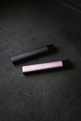 Two slim, rectangular vape devices lie on a textured dark surface. One device is dark gray and the other is pink, with the brand name visible on their sides.