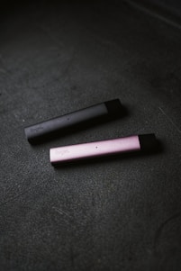 Two slim, rectangular vape devices lie on a textured dark surface. One device is dark gray and the other is pink, with the brand name visible on their sides.