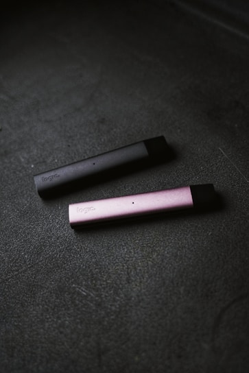 Two slim, rectangular vape devices lie on a textured dark surface. One device is dark gray and the other is pink, with the brand name visible on their sides.