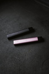 Two slim, rectangular vape devices lie on a textured dark surface. One device is dark gray and the other is pink, with the brand name visible on their sides.