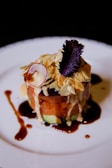 A gourmet dish artfully plated on a white ceramic plate. The dish features a stacked arrangement of sliced vegetables and raw fish or meat, topped with sliced almonds, a small red radish slice, and a dark leafy garnish. Rich sauces are drizzled around creating an artistic presentation.