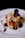 A gourmet dish artfully plated on a white ceramic plate. The dish features a stacked arrangement of sliced vegetables and raw fish or meat, topped with sliced almonds, a small red radish slice, and a dark leafy garnish. Rich sauces are drizzled around creating an artistic presentation.