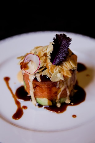 A gourmet dish artfully plated on a white ceramic plate. The dish features a stacked arrangement of sliced vegetables and raw fish or meat, topped with sliced almonds, a small red radish slice, and a dark leafy garnish. Rich sauces are drizzled around creating an artistic presentation.