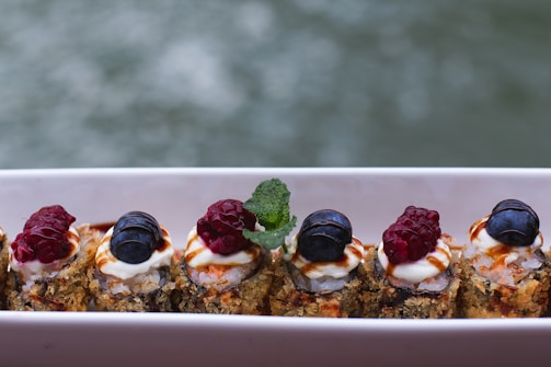A row of sushi rolls is topped with fresh raspberries and blueberries, along with a generous dollop of cream and a drizzle of sauce. A mint leaf adds a touch of green, providing contrast against the vibrant berries. The rolls sit on a rectangular white plate against a blurred background.