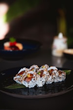 A plate of sushi rolls garnished with sesame seeds is elegantly arranged on a dark plate. The rolls are filled with a mix of ingredients, including vibrant green avocado and red fish. The background includes a blurred setting, suggesting a dining or restaurant environment.
