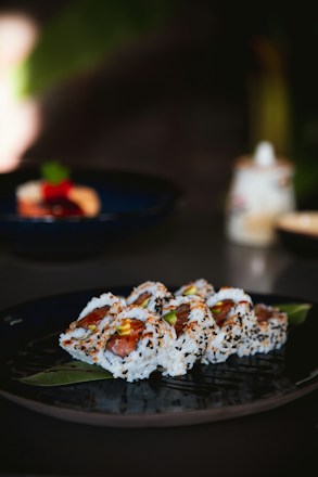 A plate of sushi rolls garnished with sesame seeds is elegantly arranged on a dark plate. The rolls are filled with a mix of ingredients, including vibrant green avocado and red fish. The background includes a blurred setting, suggesting a dining or restaurant environment.