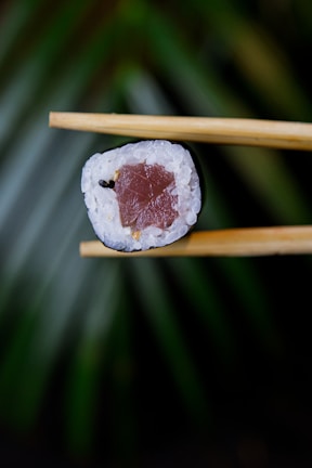 A piece of sushi roll is held delicately between two wooden chopsticks. The sushi features a center filling that appears to be raw fish, surrounded by white rice and a wrap of seaweed. The background is blurred with green tones, possibly suggesting a nature-inspired setting.
