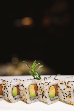 A close-up view of sushi rolls decorated with sesame seeds, featuring avocado and a slice of fish within the rice. A small green garnish is placed on top, providing a touch of freshness. The background is blurred, emphasizing the sushi.