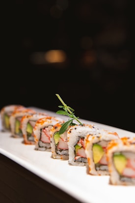 A neatly arranged row of sushi rolls lies on a white ceramic plate. Each piece showcases a filling of avocado and crab stick, wrapped in rice and sprinkled with sesame seeds. A small sprig of greenery tops the central roll, adding a touch of freshness.